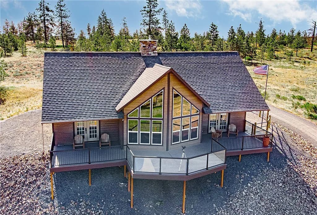 Custom home on a hillside with a wraparound deck, glass railings, and a gabled wall of windows overlooking pines
