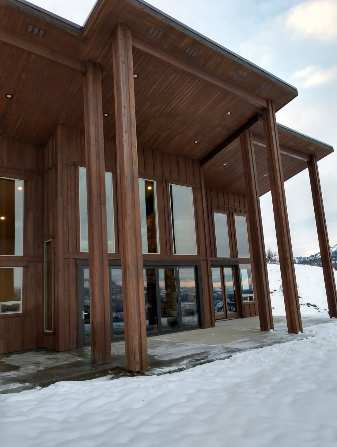 Winter view of a custom home with timber columns, vertical wood siding, and floor-to-ceiling glass
