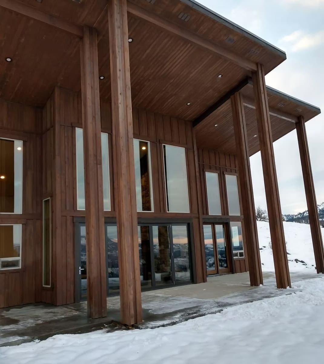 Custom timber home with a deep covered pavilion, tall wood-framed windows, and snow on the ground